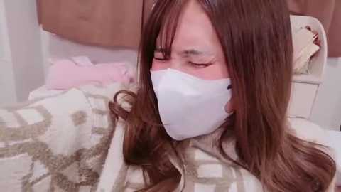 Video of a young Asian woman with long brown hair, wearing a white face mask, lying on a bed with a beige and white patterned comforter in a softly lit bedroom.