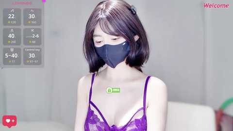 Video of an East Asian woman with straight black hair, wearing a purple lace bra, a black face mask, and a green medical tag. The background features a light-colored room with a \"Welcome\" overlay and a digital thermometer.