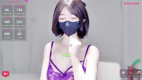 A young Asian woman with straight, shoulder-length dark hair, wearing a purple lace bra, black face mask, and green hand sanitizer, sits indoors. The background is blurred with social media icons.