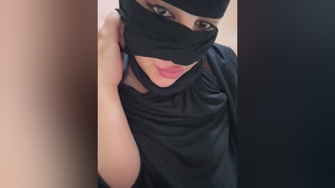 Video of a woman in a black niqab, partially covering her face, with her right hand touching her face, wearing a black headscarf.