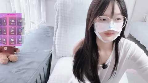 Video of an East Asian woman with long black hair and glasses, wearing a white mask, sitting on a hospital bed. The background shows a purple digital screen displaying medical data and a teddy bear.