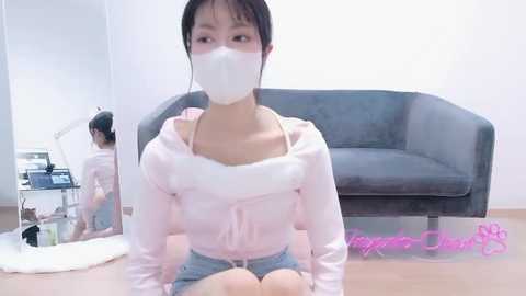 Video of an East Asian woman with fair skin, wearing a white face mask, pink hoodie, and denim shorts, kneeling on a light wood floor in a modern living room with a gray sofa and white lamp.