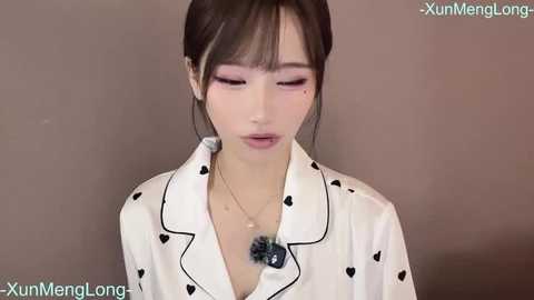 Video of an East Asian woman with light skin, dark hair in a bun, wearing a white robe with black heart patterns, a necklace, and pink makeup, against a plain background.