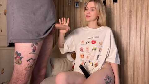Video of a blonde woman in a white graphic T-shirt, sitting on a beige couch, gesturing with her right hand, while a man in grey shorts stands in the background.