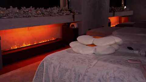 Media: Video of a dimly lit spa room with a cozy ambiance. A bed with white linens is adorned with neatly stacked pillows. A glowing fireplace and soft, warm lighting create a serene atmosphere.