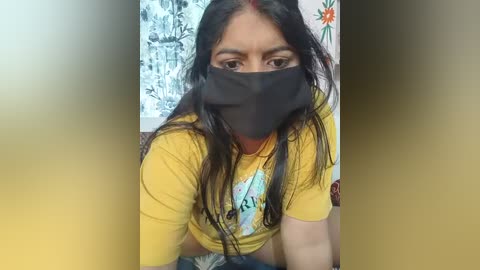 Video of a South Asian woman with long black hair, wearing a yellow t-shirt with a graphic design, black face mask, and floral wallpaper in the background.