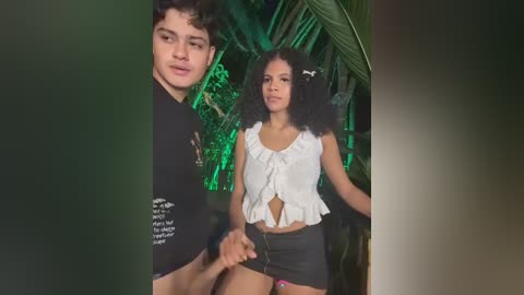 Video of a young mixed-race couple in a tropical setting; the man wears a black t-shirt, the woman a white crop top and black shorts, surrounded by lush green foliage.