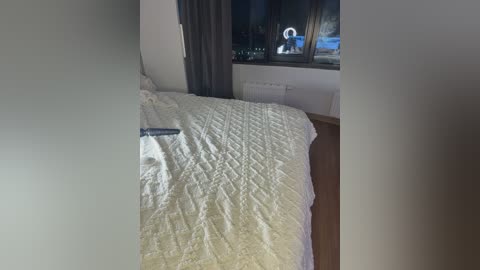 A video of a tidy, unmade bed with a white quilt, a remote control, and a window with a night view. The room has a wooden floor and light-colored walls.