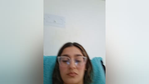 Video of a young woman with fair skin, wearing glasses, and brown hair, sitting against a white wall with a teal cushion, slightly out of focus.