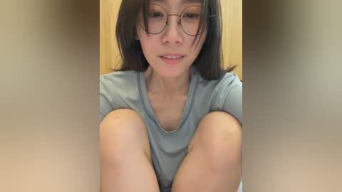 A video of an Asian woman with light skin, straight black hair, and glasses, sitting with legs bent, wearing a light blue shirt. She has a neutral expression. Background is a beige wall.