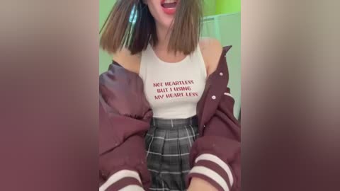 Media: A video of a young woman with straight brown hair, wearing a white tank top with a red heart graphic, a plaid skirt, and a maroon varsity jacket, smiling in a brightly lit room.