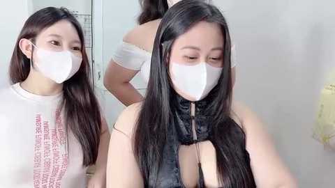 Video of an Asian woman with long, straight black hair wearing a white face mask and a sleeveless black top with a cutout. She is standing in a bright, minimalistic room with a mirror reflecting her back.