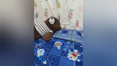 Video of a cozy child's bedroom with a brown teddy bear in a striped shirt on a blue bedspread featuring cartoon characters, against a white pillowcase with pineapple patterns.