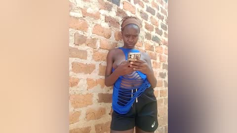 Video of a slender, dark-skinned woman with a light brown headscarf, wearing a blue, cut-out tank top and black shorts, standing against a textured, light brown brick wall, holding a smartphone.