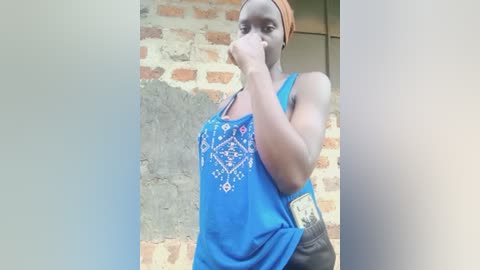 Video of a dark-skinned woman with a headscarf, wearing a blue tank top with intricate white embroidery, standing against a weathered brick wall. She holds her hand to her mouth, with a serious expression.