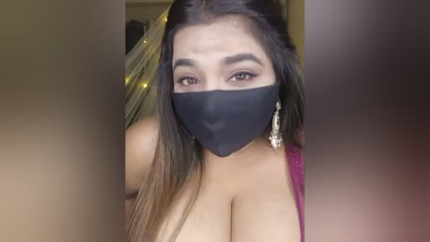 Video of a woman with long black hair, brown eyes, wearing a black mask, and a pink top, with visible cleavage.