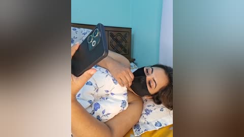 Video of a light-skinned woman with medium-length brown hair, wearing a floral sleeveless top, black mask, and a smartphone, lying on a bed with blue floral sheets and a wooden headboard, in a turquoise-painted room.