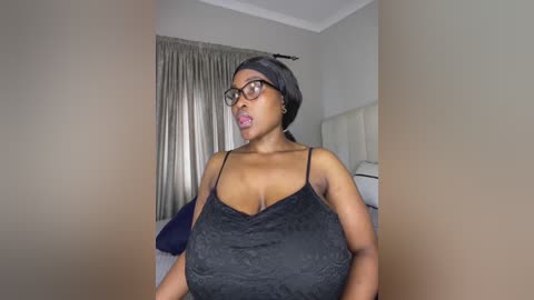 Video of a dark-skinned woman with a slender build, wearing black lace lingerie, glasses, and a black headband. She stands in a bedroom with beige walls, a bed, and curtains.