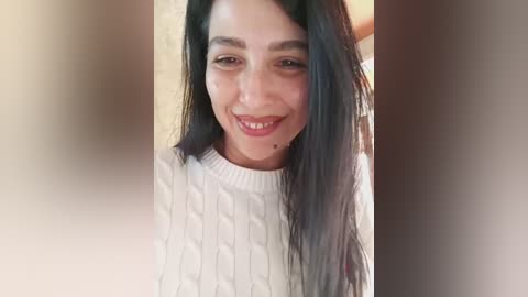 A video of a smiling, dark-haired woman with olive skin, wearing a white cable-knit sweater. The background is blurred, focusing attention on her.