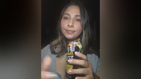 A video of a young woman with light brown skin, long dark hair, and a casual outfit, holding a colorful can of Red Bull in a dimly lit room.