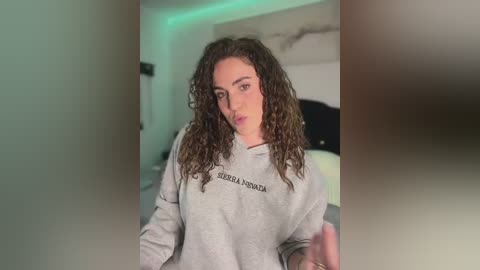 Video of a young woman with curly brown hair, light skin, wearing a gray hoodie with \"CERRA EL CIERRE\" printed on it, in a dimly lit, modern bedroom with green LED lighting.
