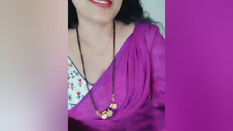 Video of a woman with fair skin, wearing a purple saree over a white blouse adorned with colorful floral embroidery. She has long, wavy black hair and is smiling, showcasing her red lipstick. A gold necklace with circular pendants hangs around her neck. The background is a gradient of pink and purple.