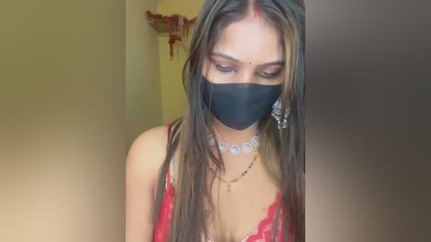 Video of a young woman with long, straight brown hair, wearing a black face mask, a red lace dress, and a blingy necklace, standing indoors against a blurred yellow wall.