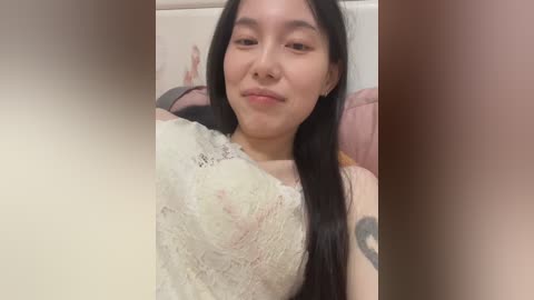Video of a young Asian woman with long black hair, wearing a white lace top, lying on a pink pillow, with a faint smile.