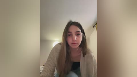 A video of a young woman with long brown hair, wearing a beige knit cardigan over a black top, standing indoors against beige walls.