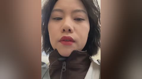 Video of a woman with medium-length black hair, light skin, and red lipstick, wearing a brown zip-up jacket, against a blurred background.