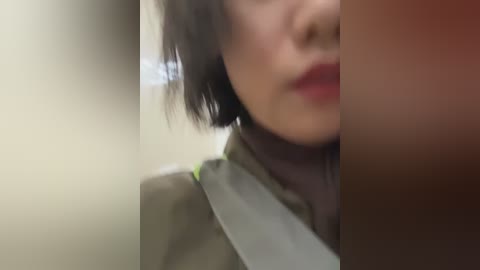 A blurry video of a person with short, dark hair, wearing a green jacket, and a seat belt across their shoulder, with a blurred background.