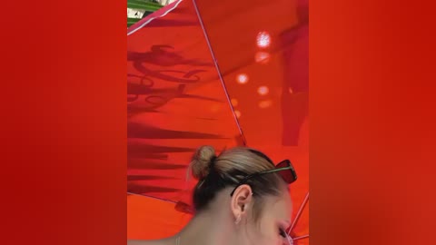 Video of a woman with blonde hair tied in a bun, wearing dark sunglasses, standing in front of a vibrant red wall with reflective surfaces. The image is overlaid with a bold red border.