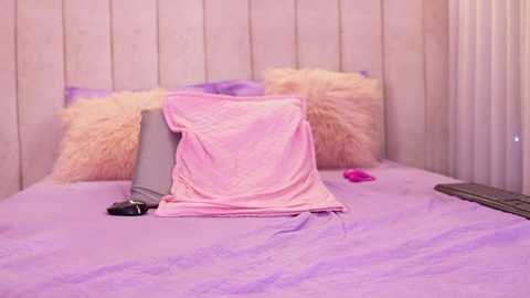 Video of a bed with a fluffy pink blanket, two fluffy pillows, and a pink toy car on a lavender bedspread, set against a light wood-paneled headboard.