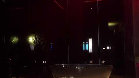 A video of a dimly-lit, modern interior with a large, round, clear glass table center. The background features dark, shadowy walls and subtle red lighting, creating an intimate, moody atmosphere.
