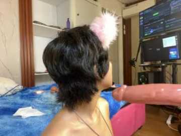 Video of a topless Asian woman with black hair, wearing pink cat ears, sitting on a blue blanket in a bedroom. A large TV screen displays an adult video.