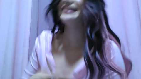 Video of a woman with long, wavy black hair, smiling, wearing a white top, in a dimly lit bathroom with a white shower curtain in the background.