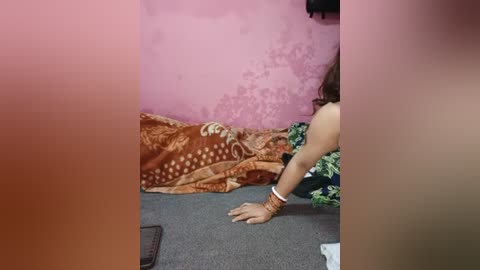 A video of a woman in a small, dimly lit room with a pink wall. She has medium skin tone and is wearing a floral-patterned dress. The floor is covered in a grey carpet, and there is a brown blanket with intricate designs on a bed in the background.