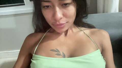 Video of a young woman with medium skin tone, long dark hair, and a small tattoo of a leaf on her chest. She wears a light green halter top and sits indoors, next to a window.