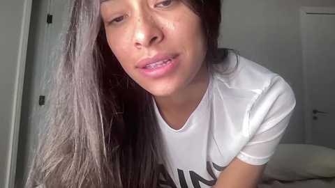 A video of a young woman with long, straight black hair, fair skin, and a slight smile, wearing a white t-shirt, taken indoors near a white door.