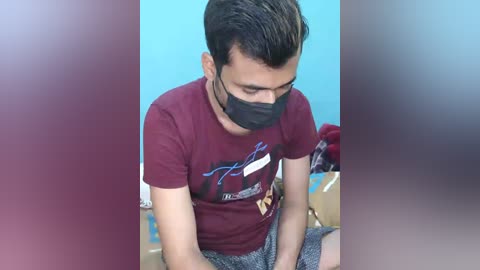 Video of a young man with short, dark hair, wearing a maroon t-shirt with a graphic, gray sweatpants, and a black face mask, seated on a bed against a blue wall, blurred background.