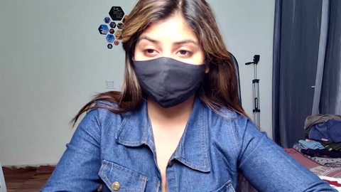 Media: Video of a woman with long brown hair, wearing a denim shirt, black mask, and black gloves, in a sparsely furnished room with a door and hexagonal patterned wall art.