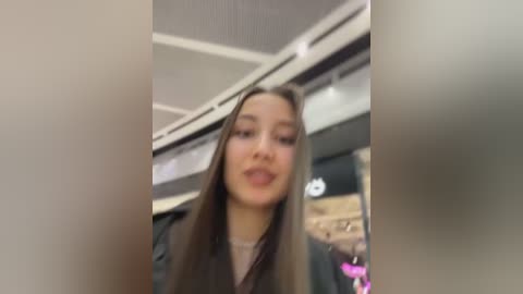 Video of a young woman with long, straight brown hair, wearing a black jacket, taken from a slightly blurry, low-angle perspective. The background features a modern, indoor setting with a ceiling fan and a pink sign.