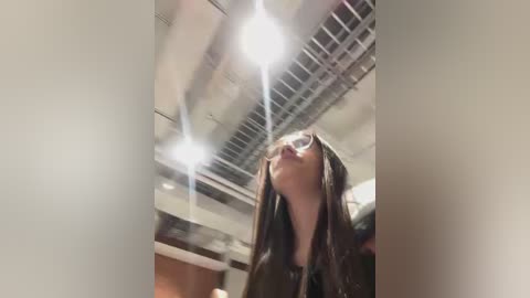 Video of a woman with long brown hair, wearing dark glasses, gazing upward at bright ceiling lights in a modern, well-lit room.