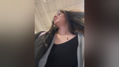 Video of a young woman with long brown hair, wearing a black top and a denim jacket, standing in a dimly lit room with exposed ceiling beams, looking upward.