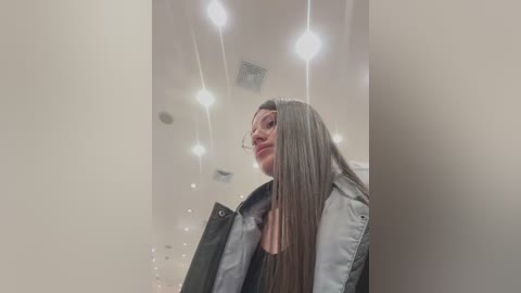 Video of a young woman with long, straight hair, wearing glasses, a black jacket, and a white shirt, standing in a modern, well-lit room with hanging light fixtures.