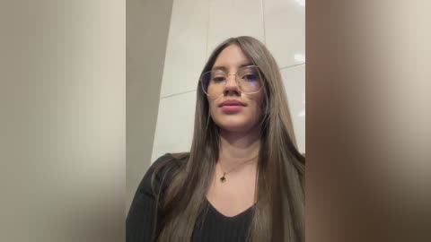 Video of a young woman with long brown hair, wearing glasses and a black top, standing in a beige-tiled room.