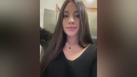 Video of a young Latina woman with long, straight dark brown hair, wearing a black top, smiling softly. She stands indoors with beige walls and a wooden door in the background.
