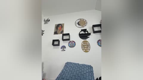 Video of a minimalist, modern room with a white wall adorned with eclectic wall art, including framed photos, clocks, and decorative plates. A blue knitted blanket covers a bed in the foreground.