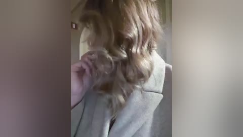 Video of a woman with wavy, light brown hair, wearing a light grey coat, standing in a dimly lit hallway. Her right hand is brushing her hair.