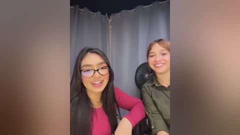 Video of two women, one with long black hair and glasses, smiling in a pink top, seated next to a woman with short brown hair and a green jacket. Background features a gray curtain.